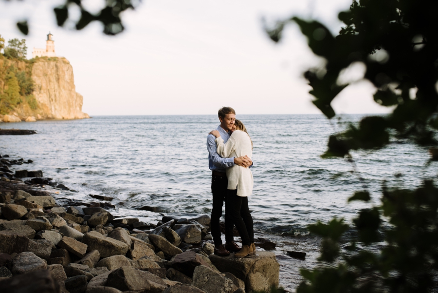 engaged couple embraces beside lake superior with lighthouse