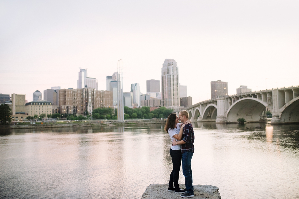 couple standing on rock beside mississippi with minneapolis behind