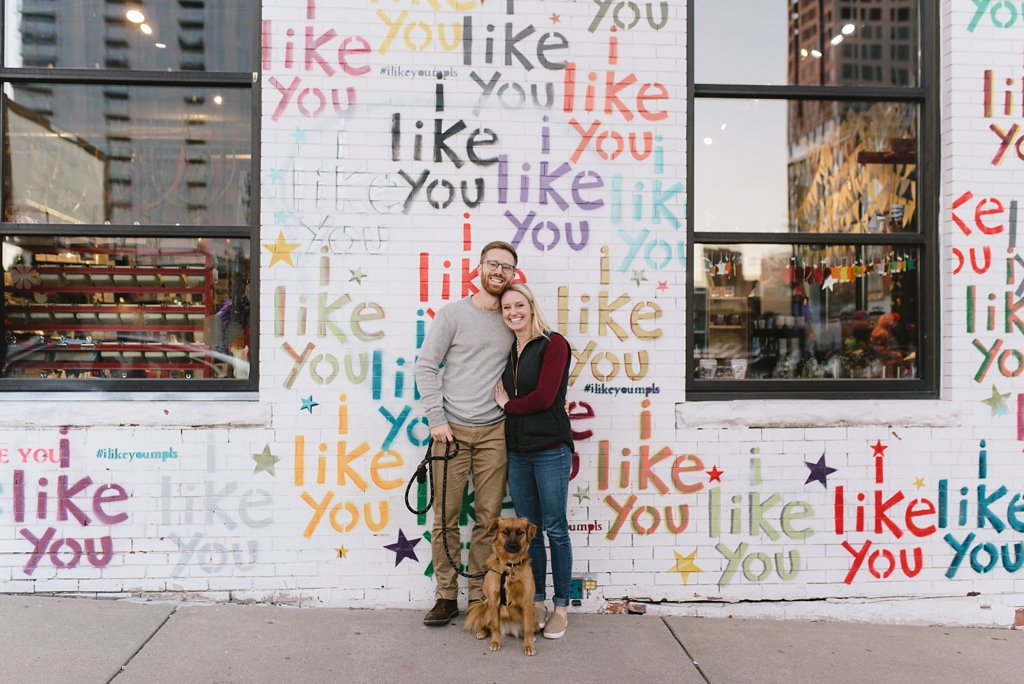 engaged couple and dog stand in front of a wall that says I like you 