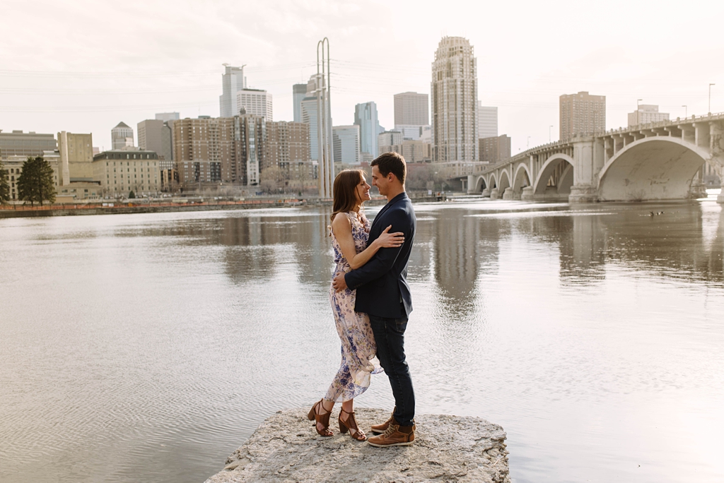 engaged couple embracing beside mississippi river with minneapolis skyline behind