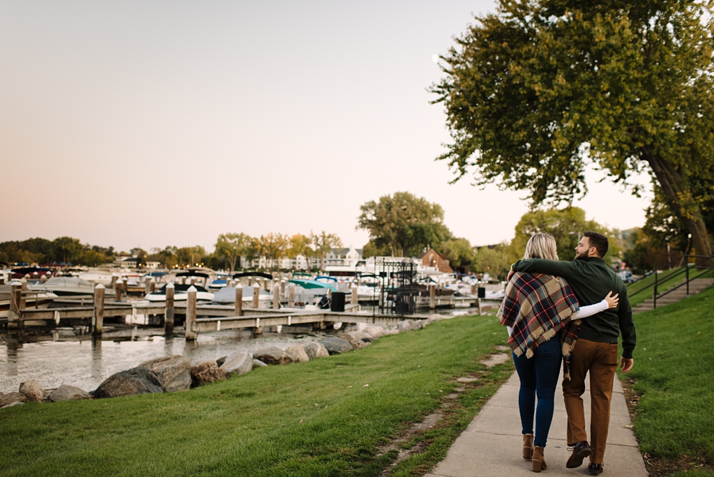engaged couple walking beside excelsior bay with boats at sunset