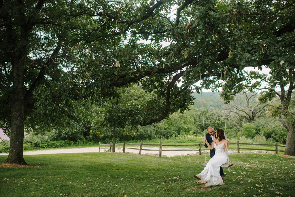 groom pushes bride on swing under large tree at winery wedding