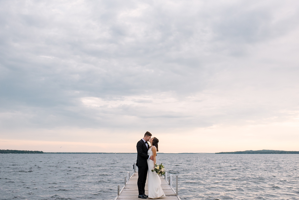 newlyweds kiss on lake dock with cloudy sky