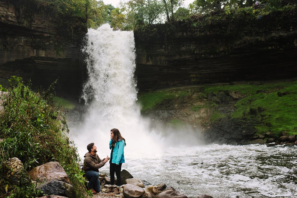 man on his knees proposing to woman in front of a waterfall