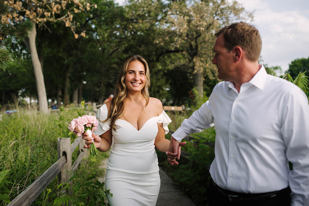 smiling bride is led down a park path by husband