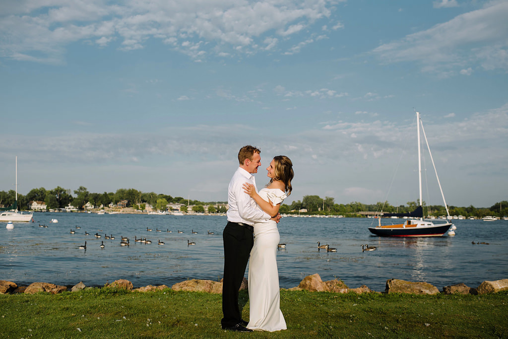 smiling newlyweds embrace in front of blue skies and lake with geese
