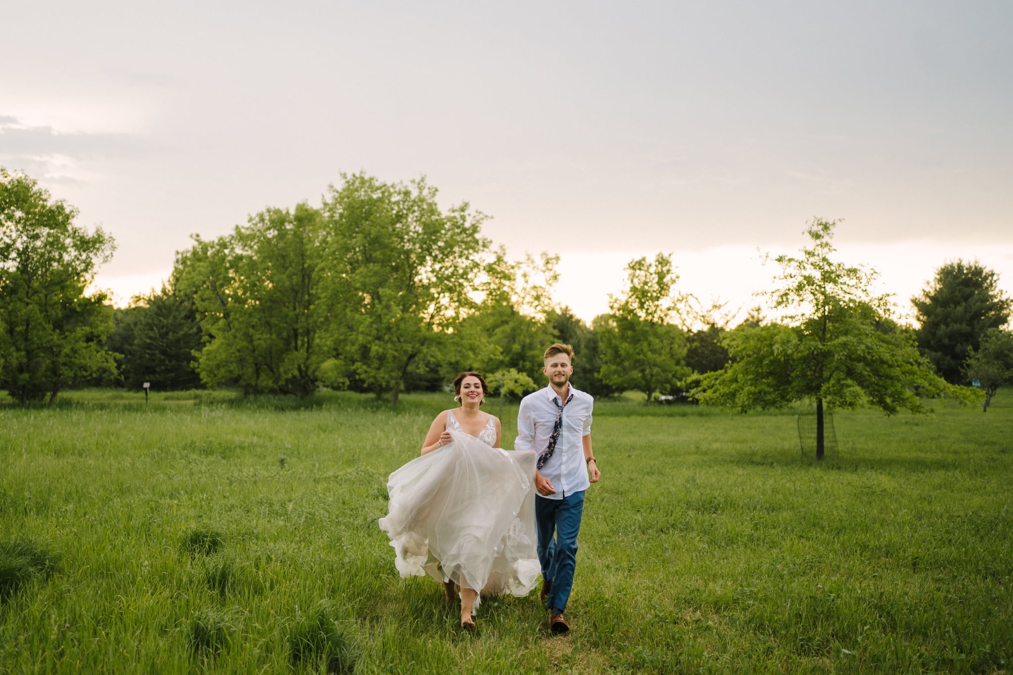 bride and groom run through green field after garden party wedding